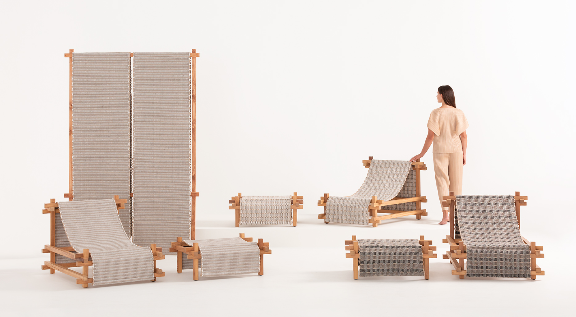 Several pieces of modern furniture with wooden frames and woven fabric surfaces arranged in a studio setting with a woman inspecting the furniture.