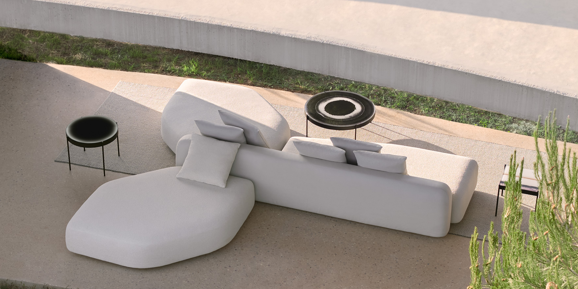 Outdoor seating area with white modern furniture, black side tables, and a textured rug on a concrete patio, with greenery nearby.