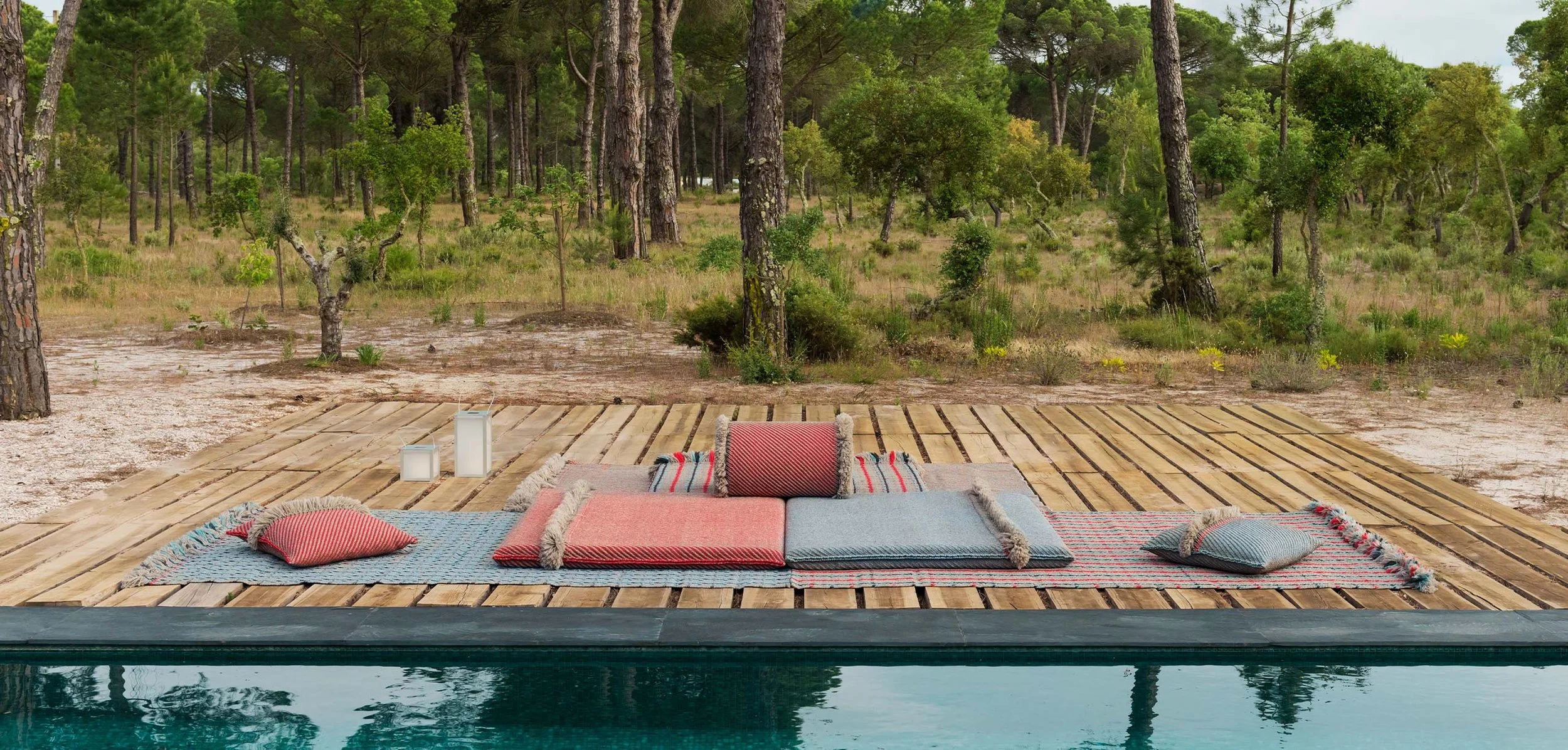 Outdoor patio with colorful cushions and blankets on a wooden deck overlooking a forest with tall trees.