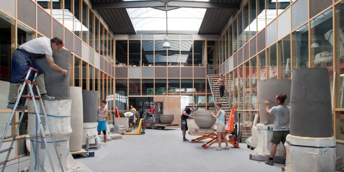 People working inside a large, well-lit workshop or studio, shaping large concrete or stone bowls, with glass walls and a high ceiling.