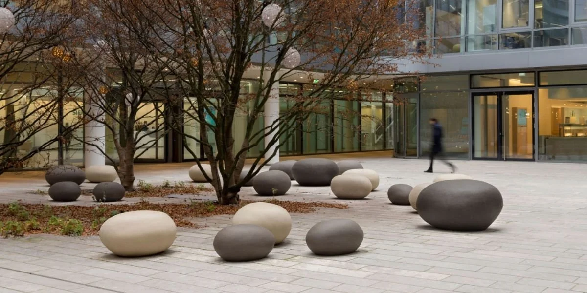 An outdoor courtyard with a leafless tree, several round rocks of varying sizes and colors, and modern glass buildings in the background. A person is walking past.