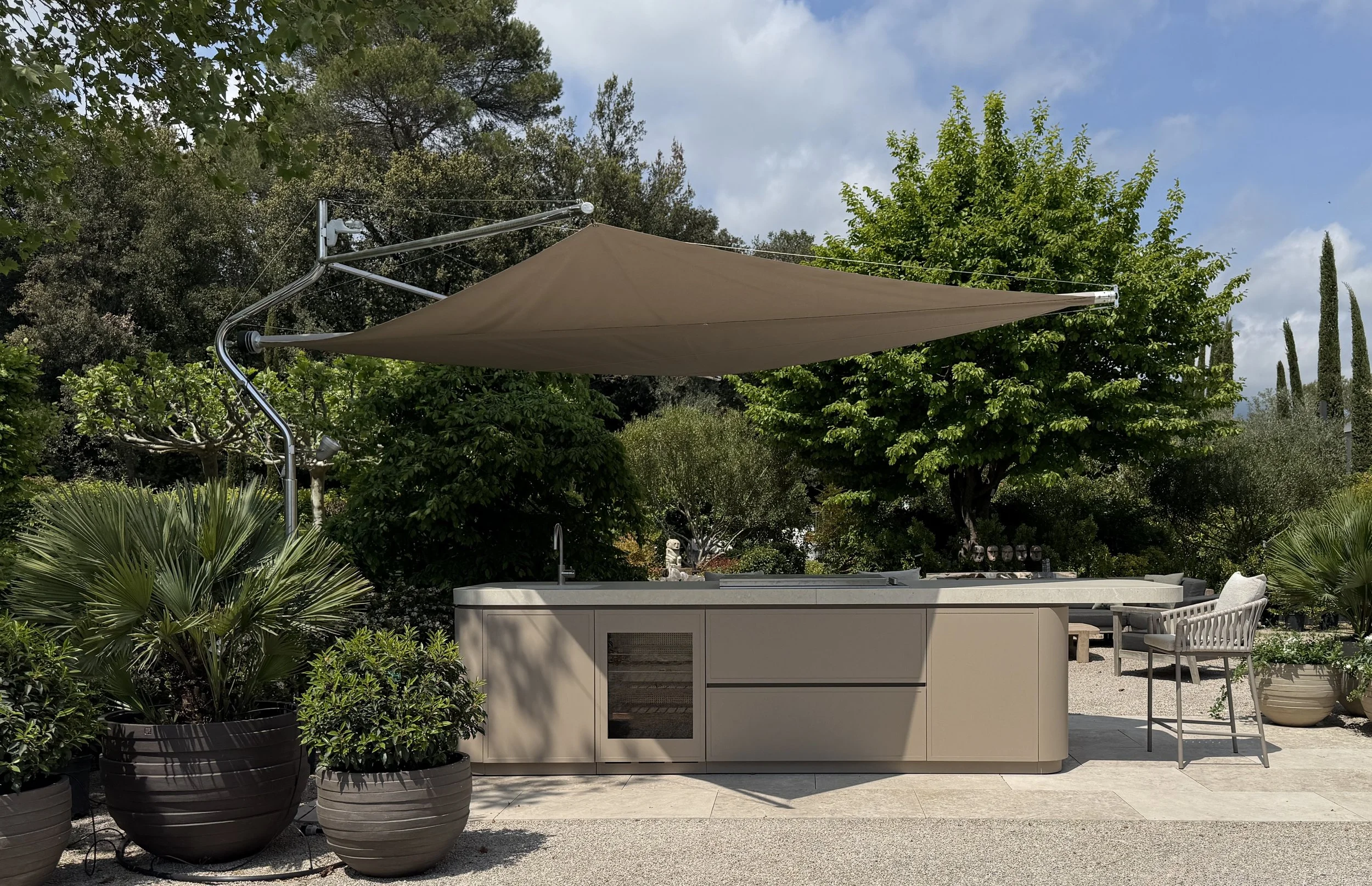 An outdoor patio with potted plants, a beige cabinet with a glass door, and a patio umbrella in a lush garden with trees and shrubs.