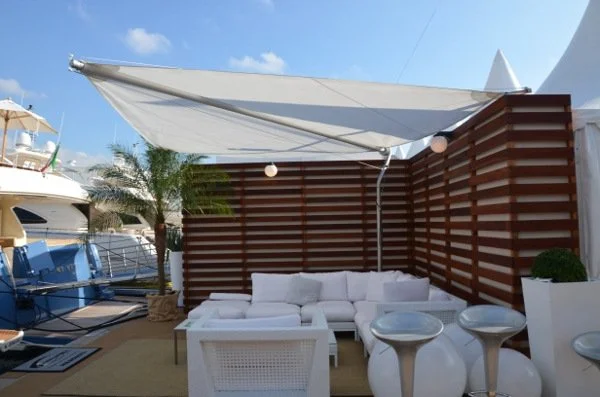 Outdoor seating area with white sofa, two white chairs, small tables, palm trees, and boats docked nearby under a blue sky.