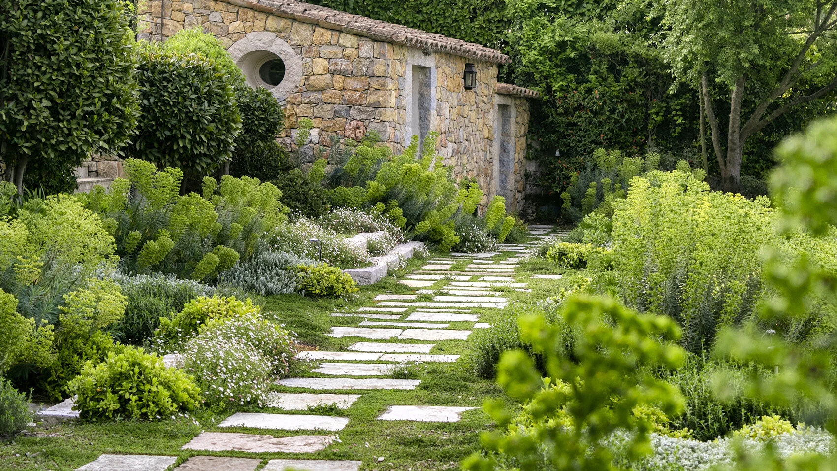 A garden path made of stone slabs winding through lush green plants and shrubs, with a rustic stone building and trees in the background.