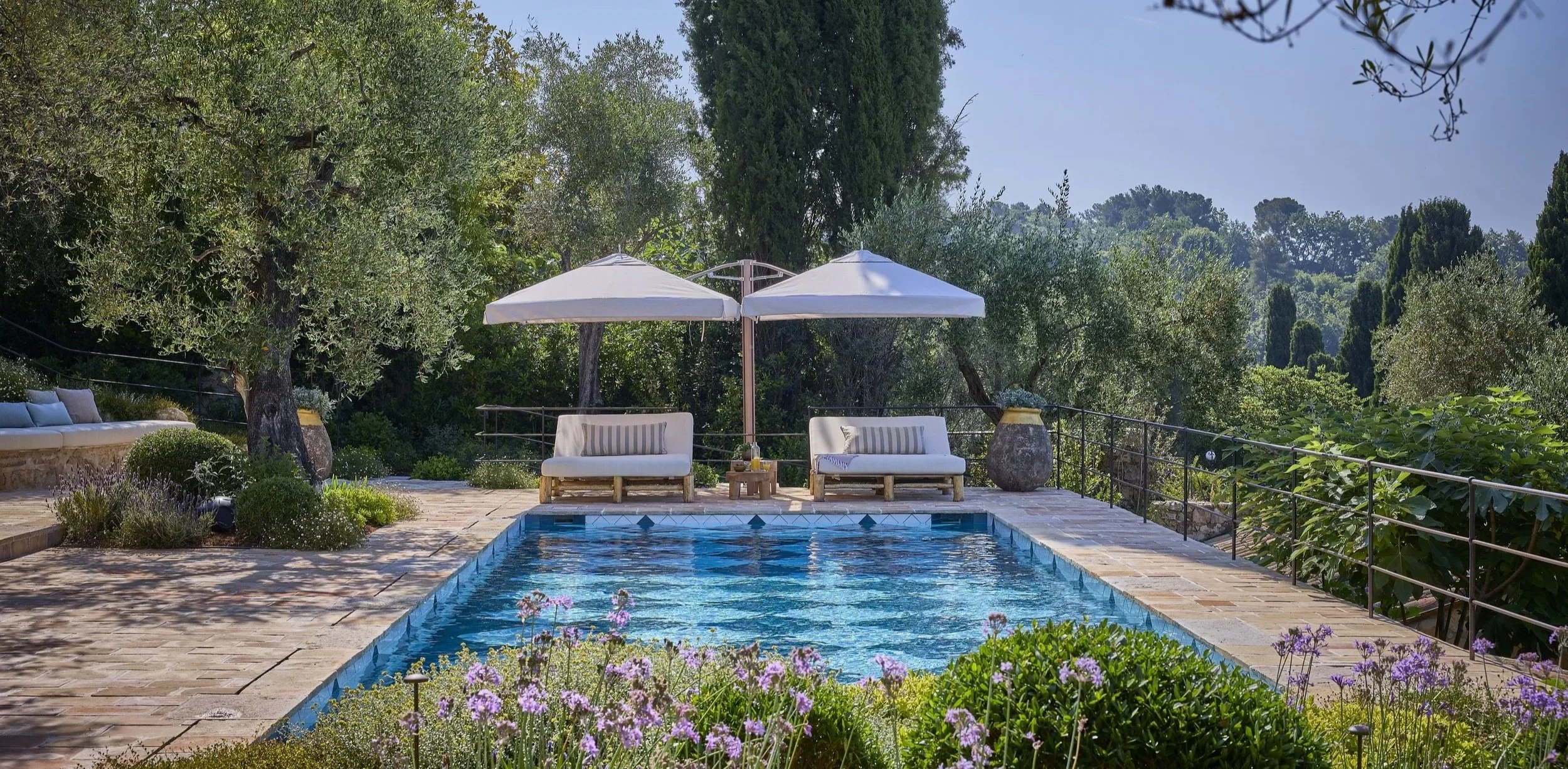 A backyard swimming pool with lounging chairs and parasols, surrounded by greenery and flowering plants.