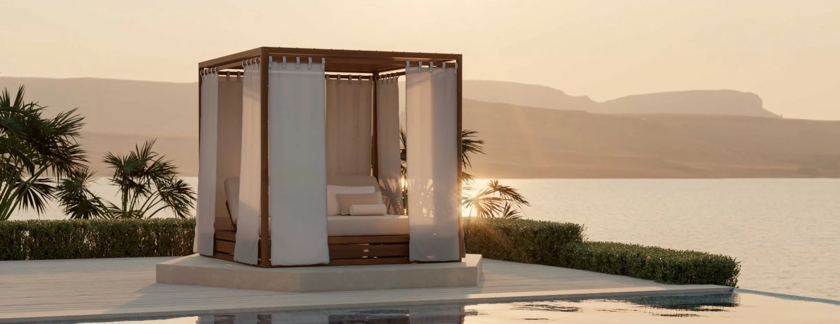 A luxurious cabana with white curtains and cushions, set by a pool and overlooking a body of water during sunset, with palm trees and distant hills in the background.