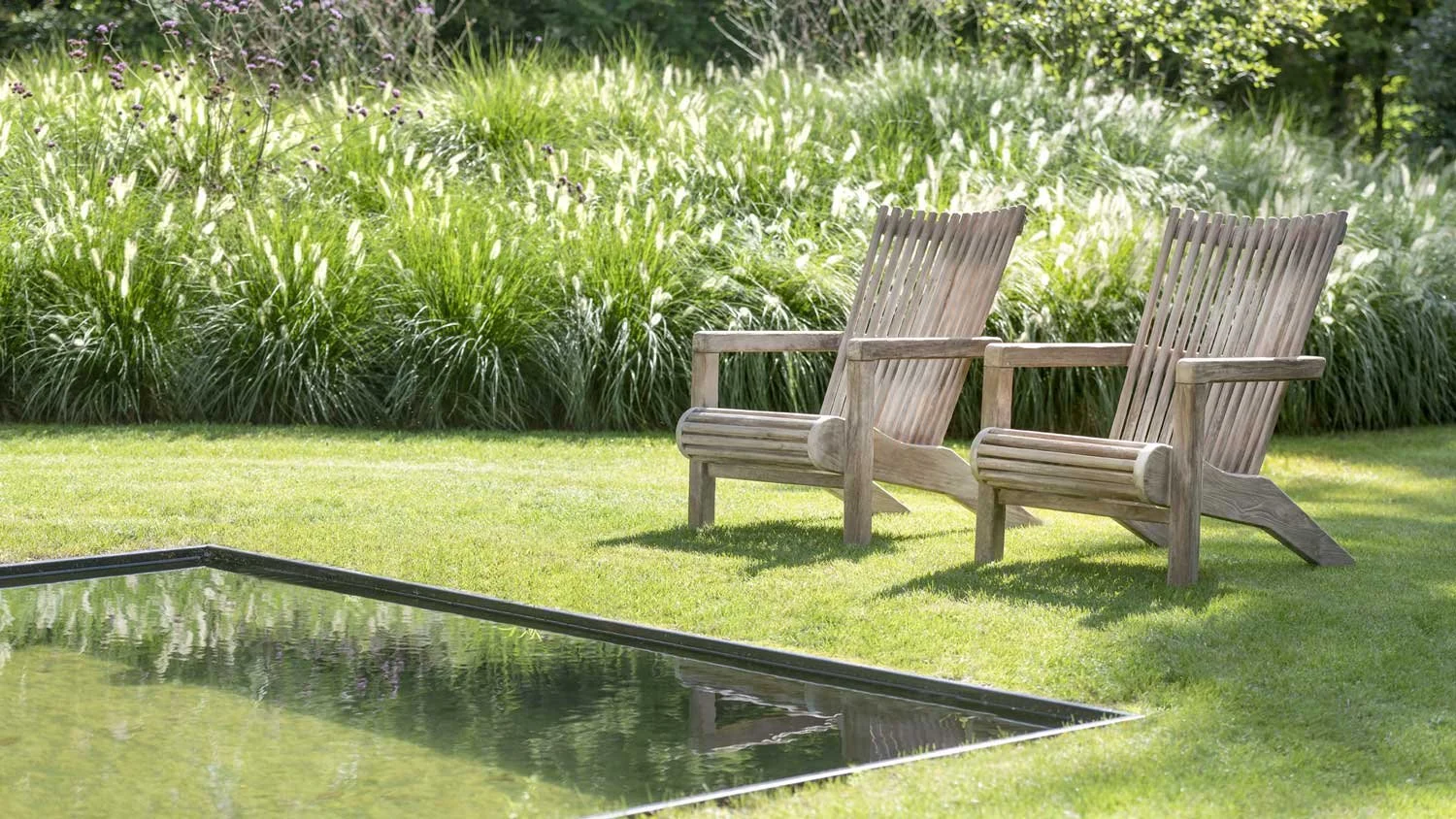Two wooden Adirondack chairs sit on a lawn near a pool, with tall ornamental grass and bushes in the background.