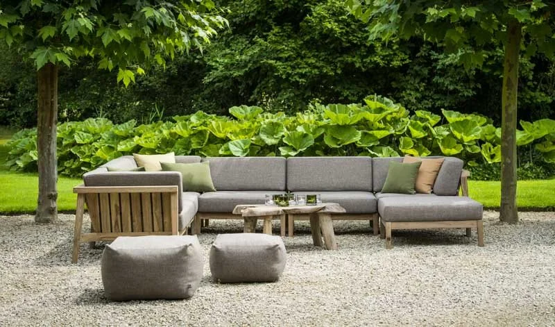 Outdoor patio furniture with a large gray sectional sofa, small poufs, and a rustic wooden coffee table on a gravel surface, surrounded by green trees and large leafy plants.