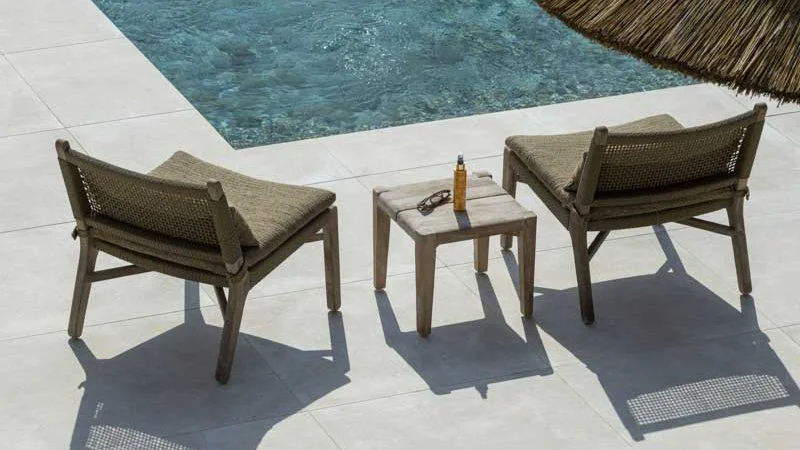 Two outdoor chairs with cushions and a small table between them, near a swimming pool with a thatched roof overhead.