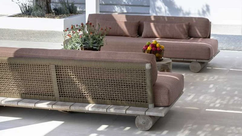 Outdoor patio area with two modern pink upholstered sofas, a wooden coffee table with a fruit bowl, and a potted plant, with sunlight casting shadows.
