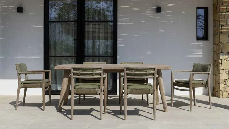 Outdoor dining table with six chairs on patio in front of a white wall and large black-framed glass doors.