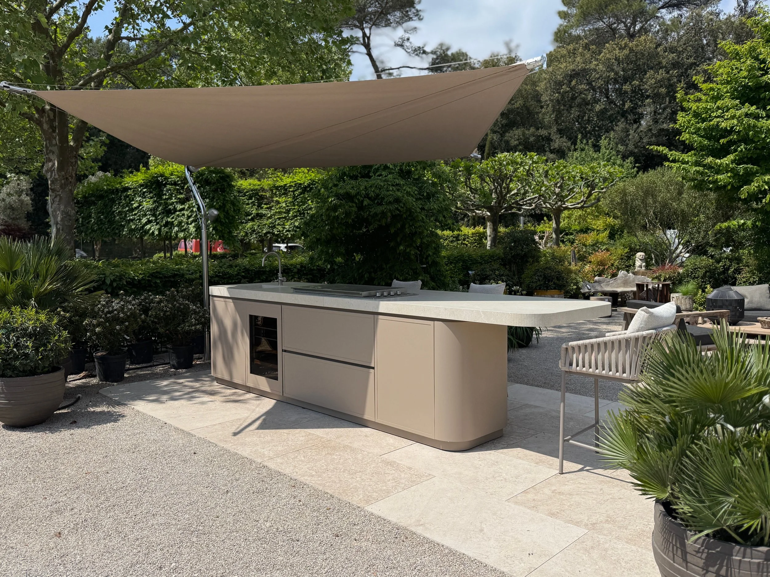 Outdoor kitchen with beige cabinetry, a sink, and a large beige shade sail overhead amidst lush green trees and plants.