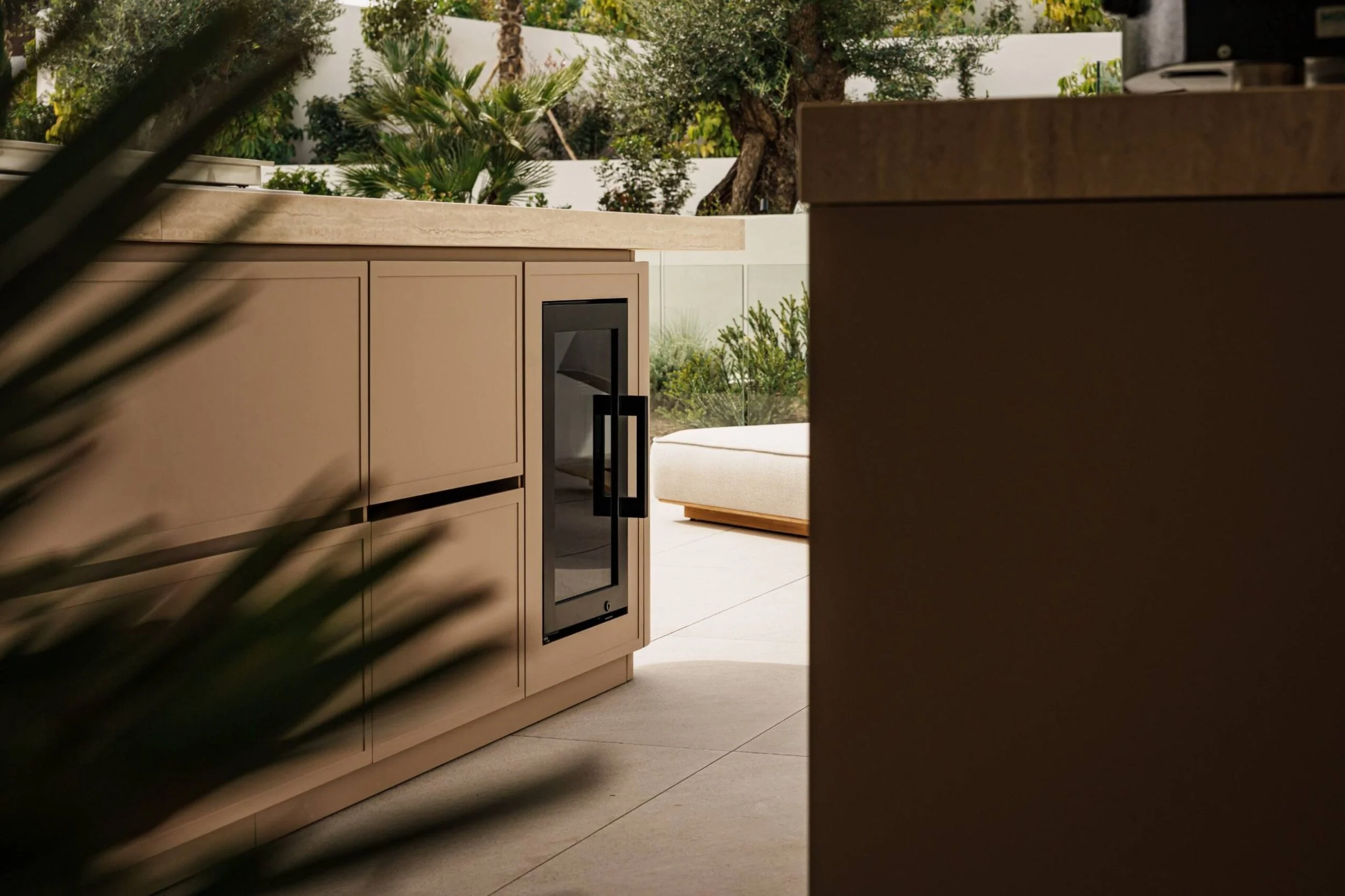Outdoor lounge area with beige cabinetry, a built-in wine fridge, and a view of lush green plants and trees.