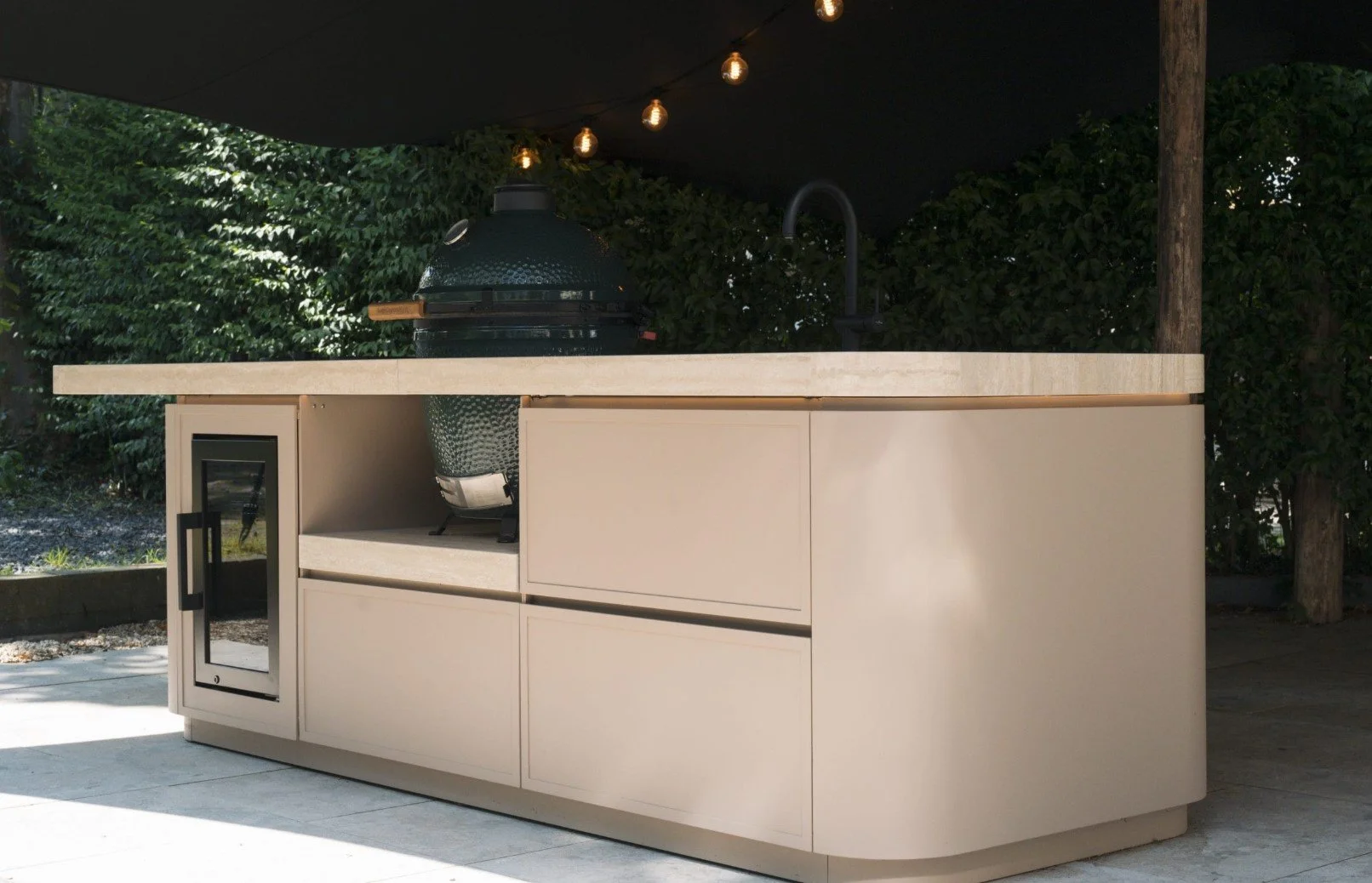 Outdoor kitchen with beige cabinetry, a built-in mini fridge, a green ceramic grill, and string lights overhead, with greenery in the background.