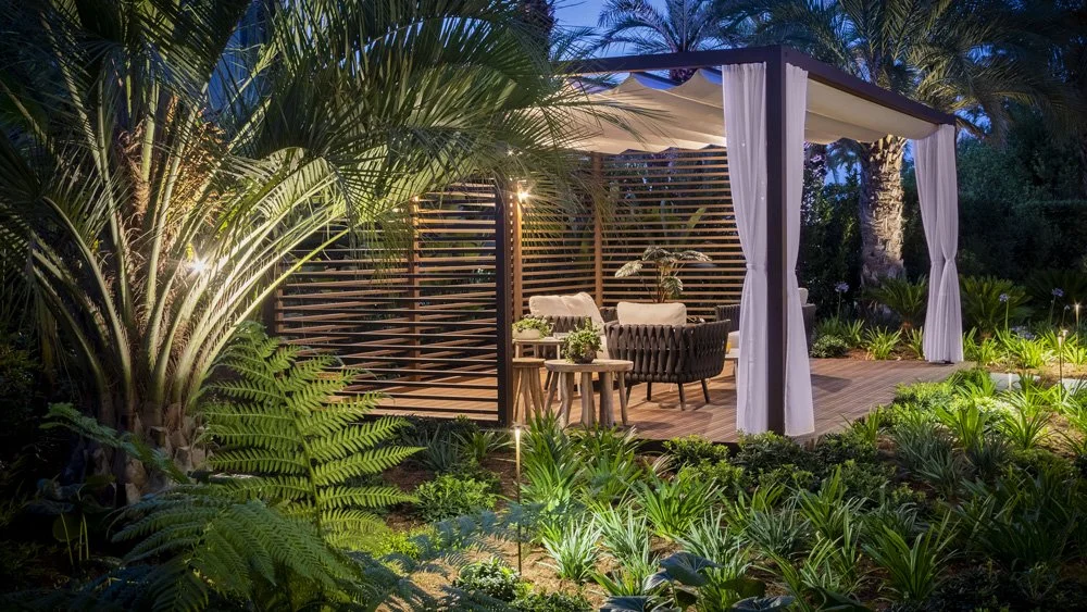 Outdoor outdoor living space with cabana, cushioned chairs, and surrounded by lush tropical plants at dusk.