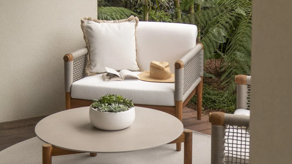 A white outdoor sofa with beige cushion, a straw hat, and an open book, accompanied by a white round coffee table with a potted succulent plant, all surrounded by green plants.