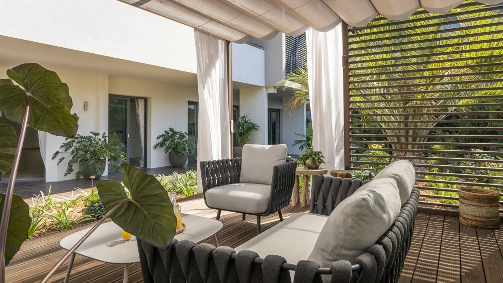 Outdoor patio with modern seating, large plants, and wooden slat divider, overlooking a garden and white building with glass doors.