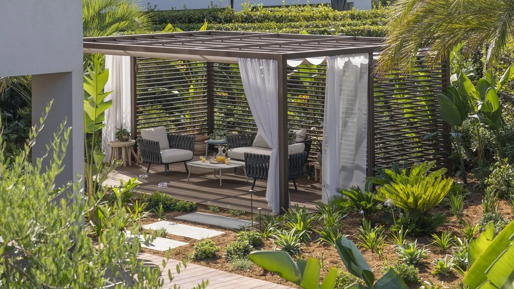 Outdoor patio with seating area, four chairs, a small coffee table with drinks, surrounded by lush green plants and wooden fencing, with white curtains hanging from a pergola.
