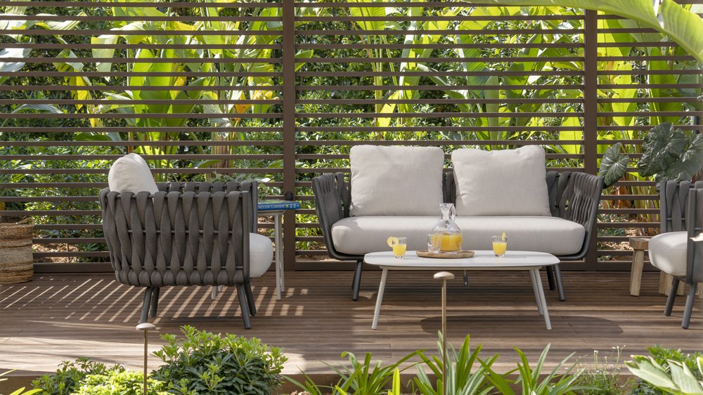 Outdoor living area with grey woven armchairs, a cream cushioned sofa, a small white coffee table with drinks, and lush green plants behind wooden slat fencing.