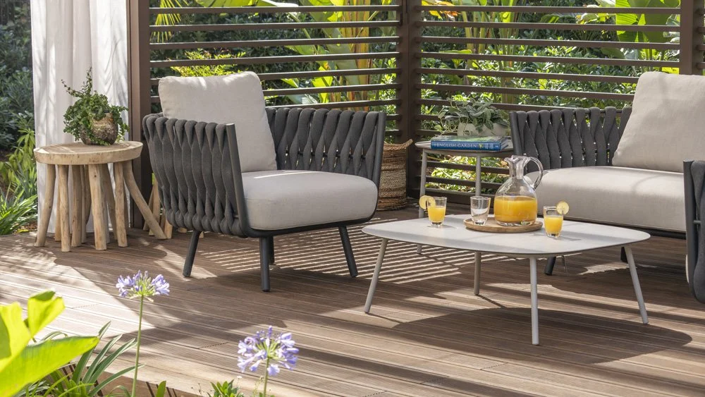 Outdoor patio with modern seating, a white coffee table with a pitcher and glasses of orange juice, surrounded by plants and wooden fencing.