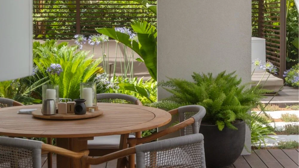 Outdoor patio with round wooden dining table, rattan chairs, potted green plants, and decorative candles, overlooking a lush garden with large leafy plants and purple flowers.
