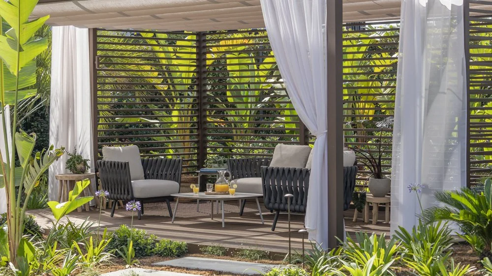 Outdoor patio with two black wicker chairs with gray cushions, a small coffee table with orange juice, surrounded by green plants and trees, with white curtains and slatted wooden privacy screens.