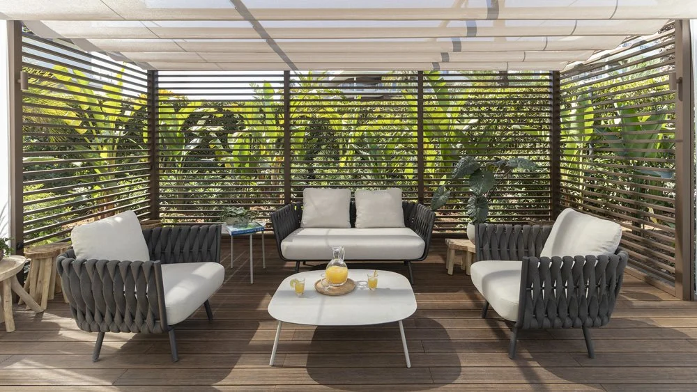 Outdoor patio with four white cushioned chairs, two black woven armchairs, a white coffee table with a pitcher and glasses, and lush green plants behind a slatted wooden privacy screen.