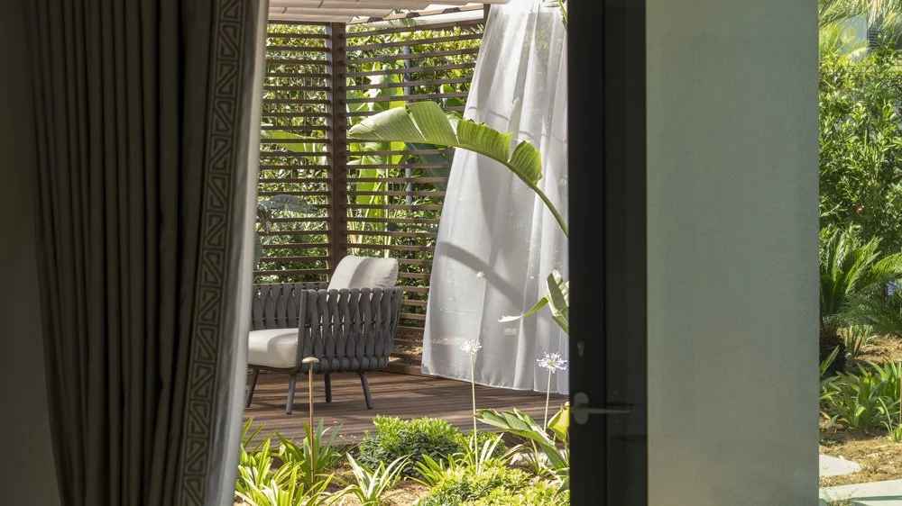 View of an outdoor patio with a beige chair, wooden slatted wall, and lush green plants, seen through a doorway with curtains.