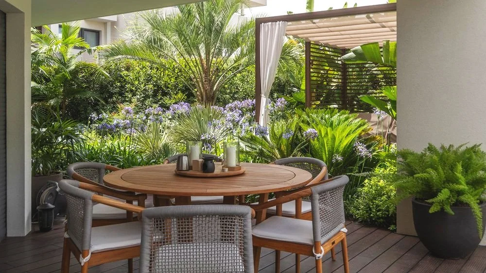 Outdoor patio with a round wooden table and six chairs surrounded by lush greenery and tropical plants.