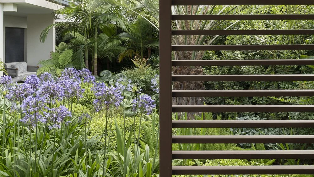 View of a lush garden with purple flowers and green foliage, partially viewed through a slatted wooden fence, with a balcony or patio area and outdoor seating in the background.