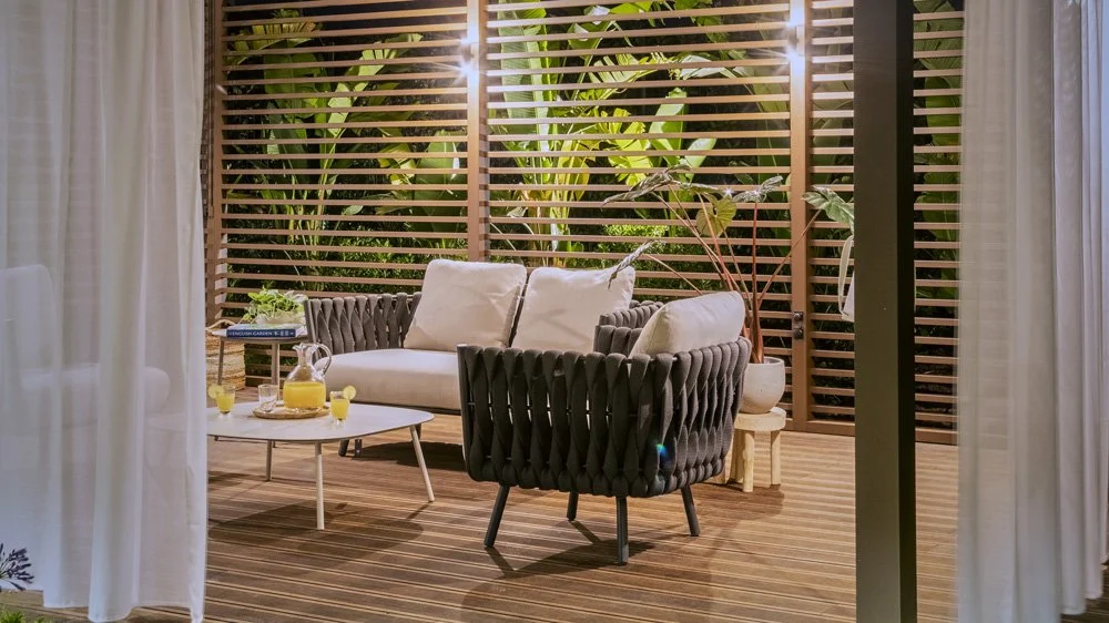 Cozy outdoor patio with black armchair, white loveseat, wooden side table, potted plants, and wooden slatted privacy screen.