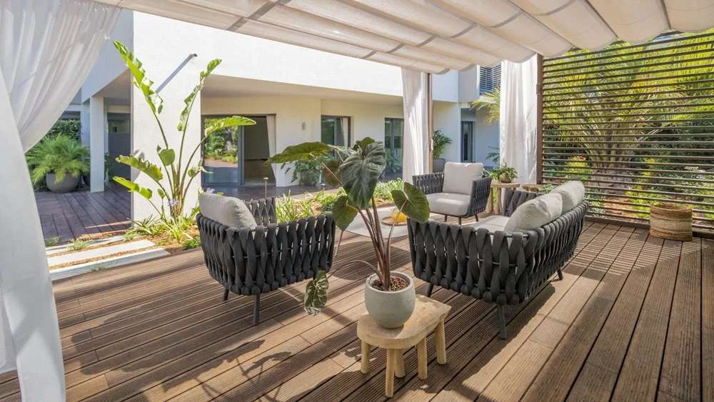 Outdoor patio area with cushioned chairs, a large potted plant, and wooden deck, surrounded by greenery and a slatted privacy wall.