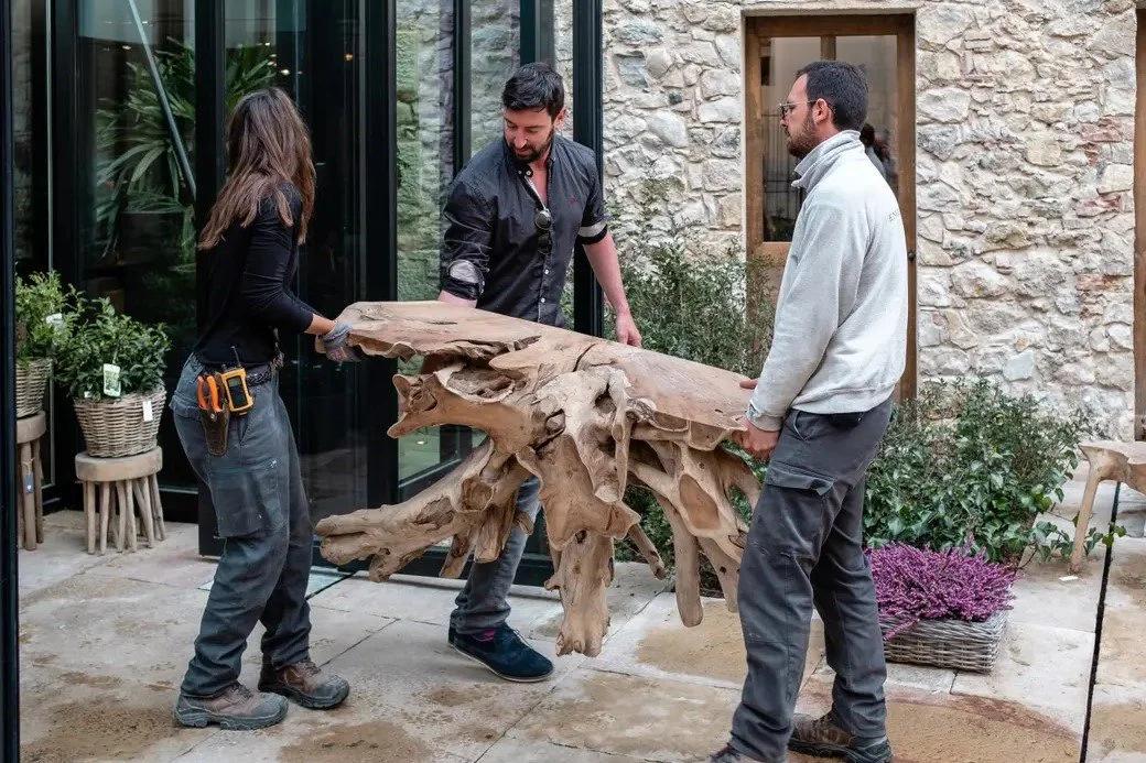 Three people are moving a large, irregularly shaped wooden sculpture outdoors near a glass building and stone wall. All are wearing work clothes.