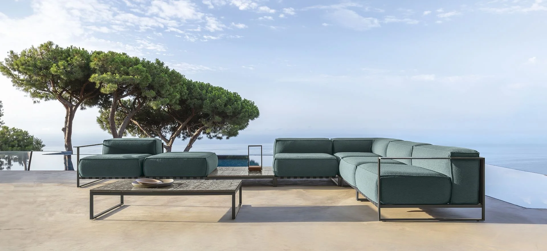 Outdoor seating area with modern teal sofas, a wooden coffee table with bowls, set on a concrete patio overlooking trees, ocean, and a partly cloudy sky.