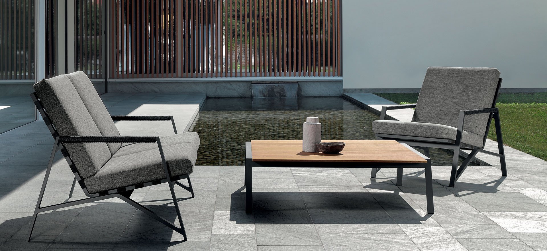 Modern outdoor patio with two gray cushioned armchairs, a wooden coffee table with vases and a bowl, a water feature, and a wooden privacy screen.