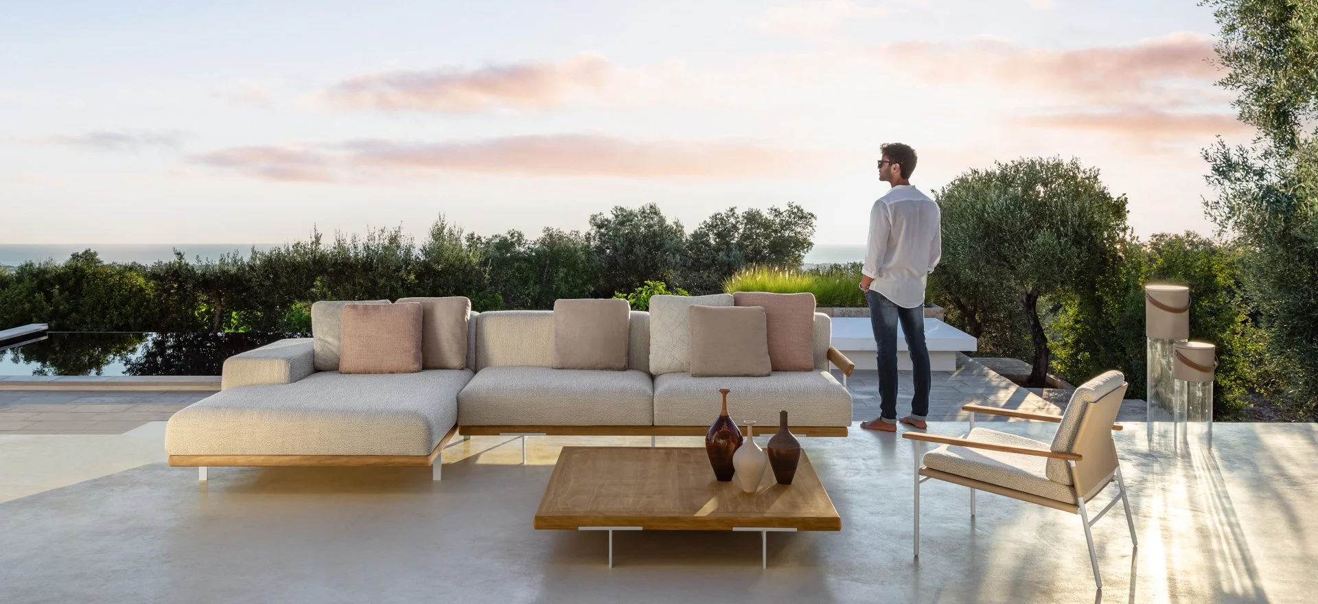 A modern outdoor patio with a beige sectional sofa, a wooden coffee table with decorative vases, a beige armchair, and a man standing near the edge looking at a scenic view of trees, a body of water, and a pastel-colored sky at sunset.