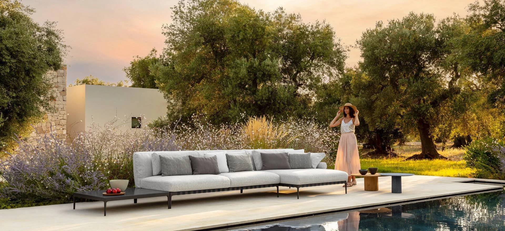 A woman standing outdoors by a pool at sunset, wearing a wide-brimmed hat and summer dress, with a white outdoor sofa with gray pillows, trees, and flowering bushes in the background.
