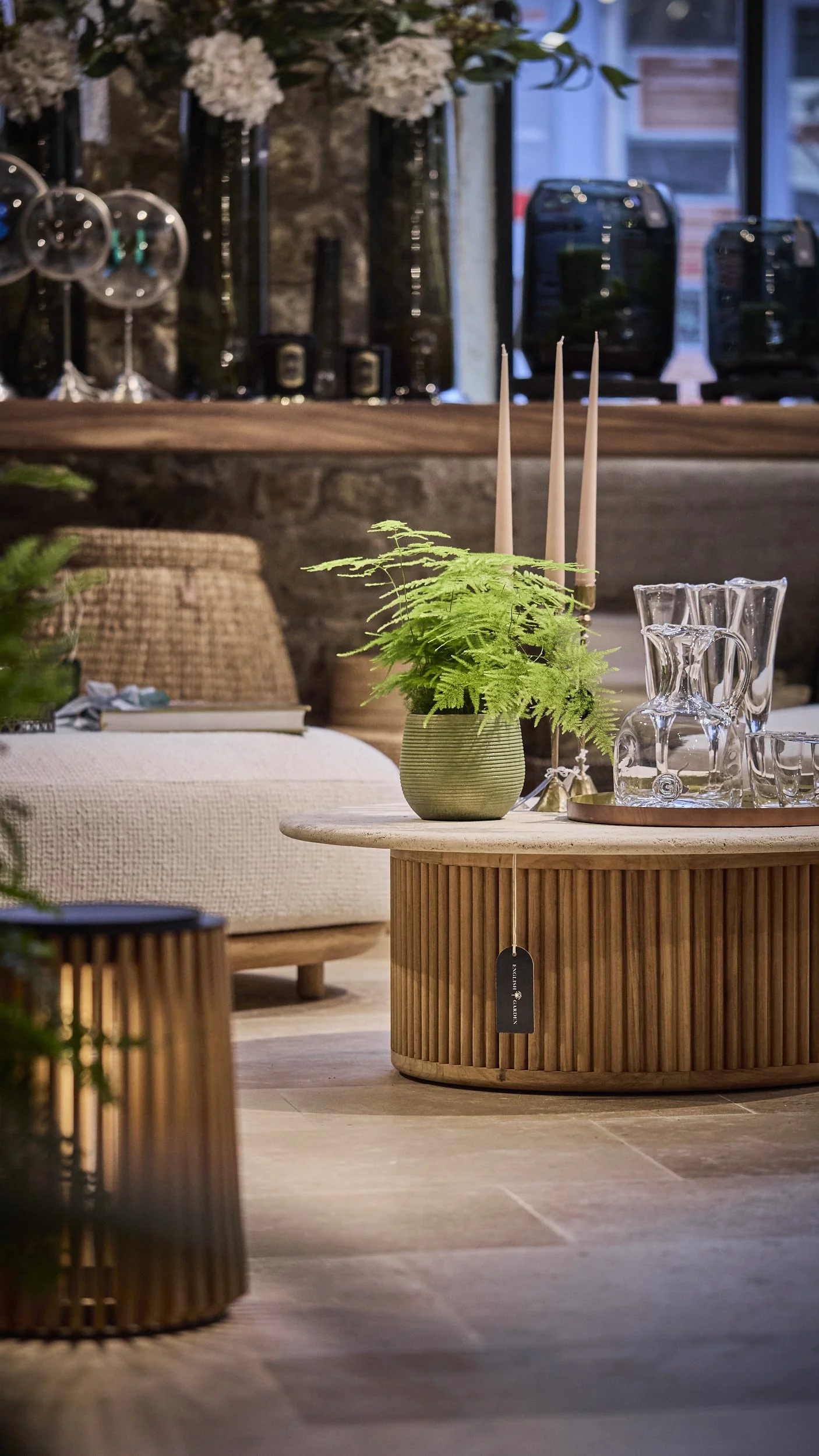 Living room with a round wooden coffee table, green potted fern, glass decanter set, and a beige sofa in the background, decorated with candles and flowers.