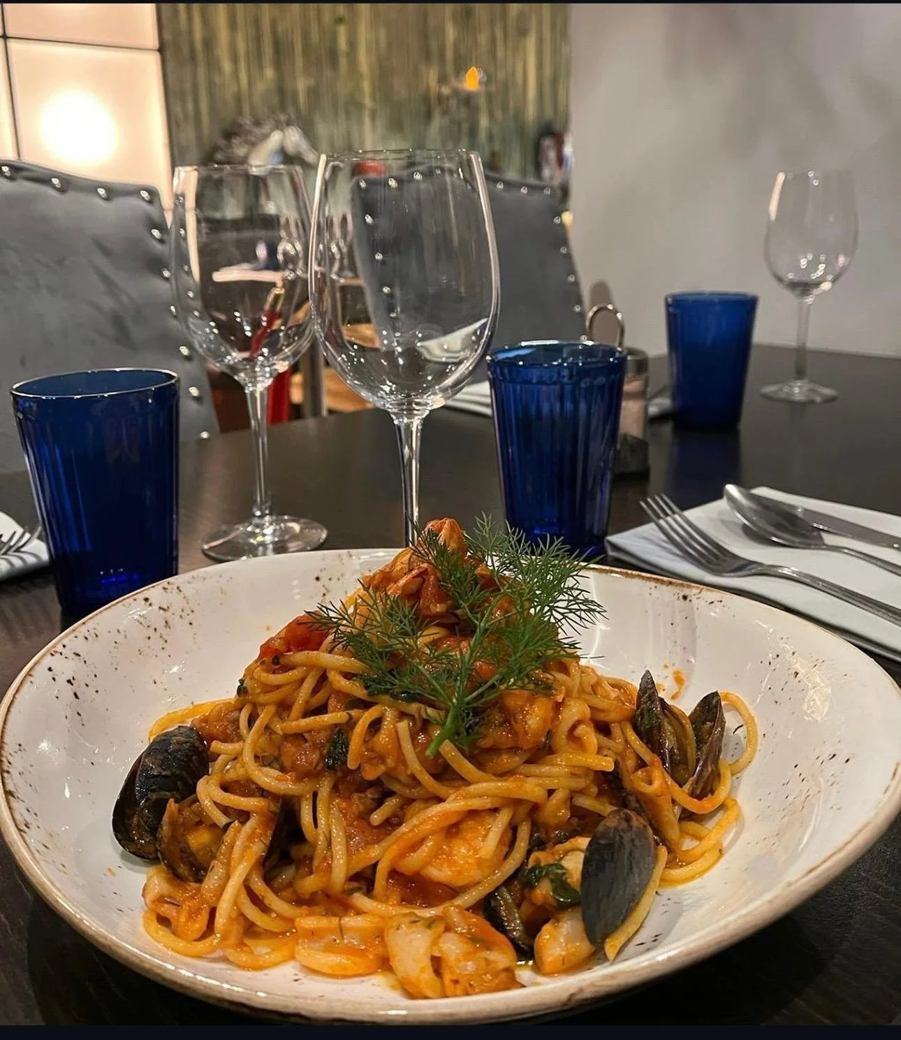 A plate of seafood pasta garnished with fresh herbs on a table set with wine glasses, blue drinking glasses, and silverware, in a restaurant setting.