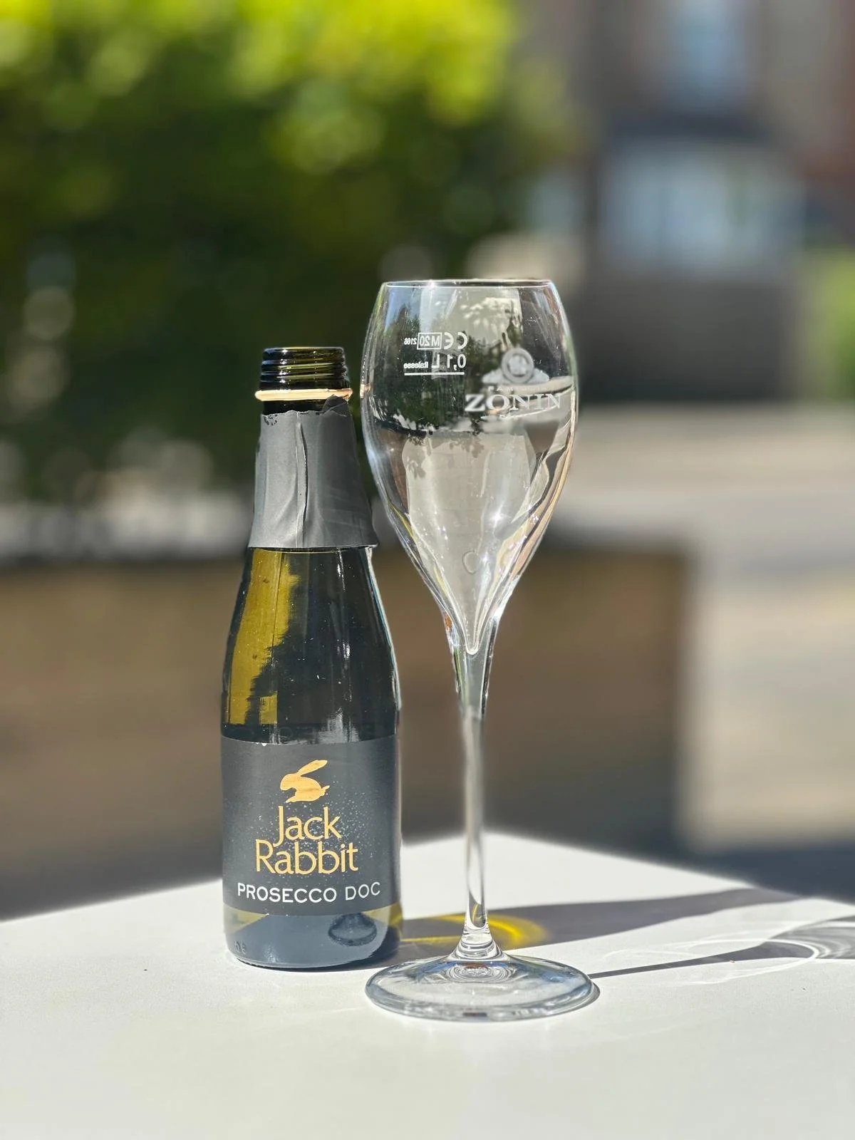 A bottle of Jack Rabbit Prosecco DOC and an empty champagne flute on a white surface outdoors with blurred green trees in background.