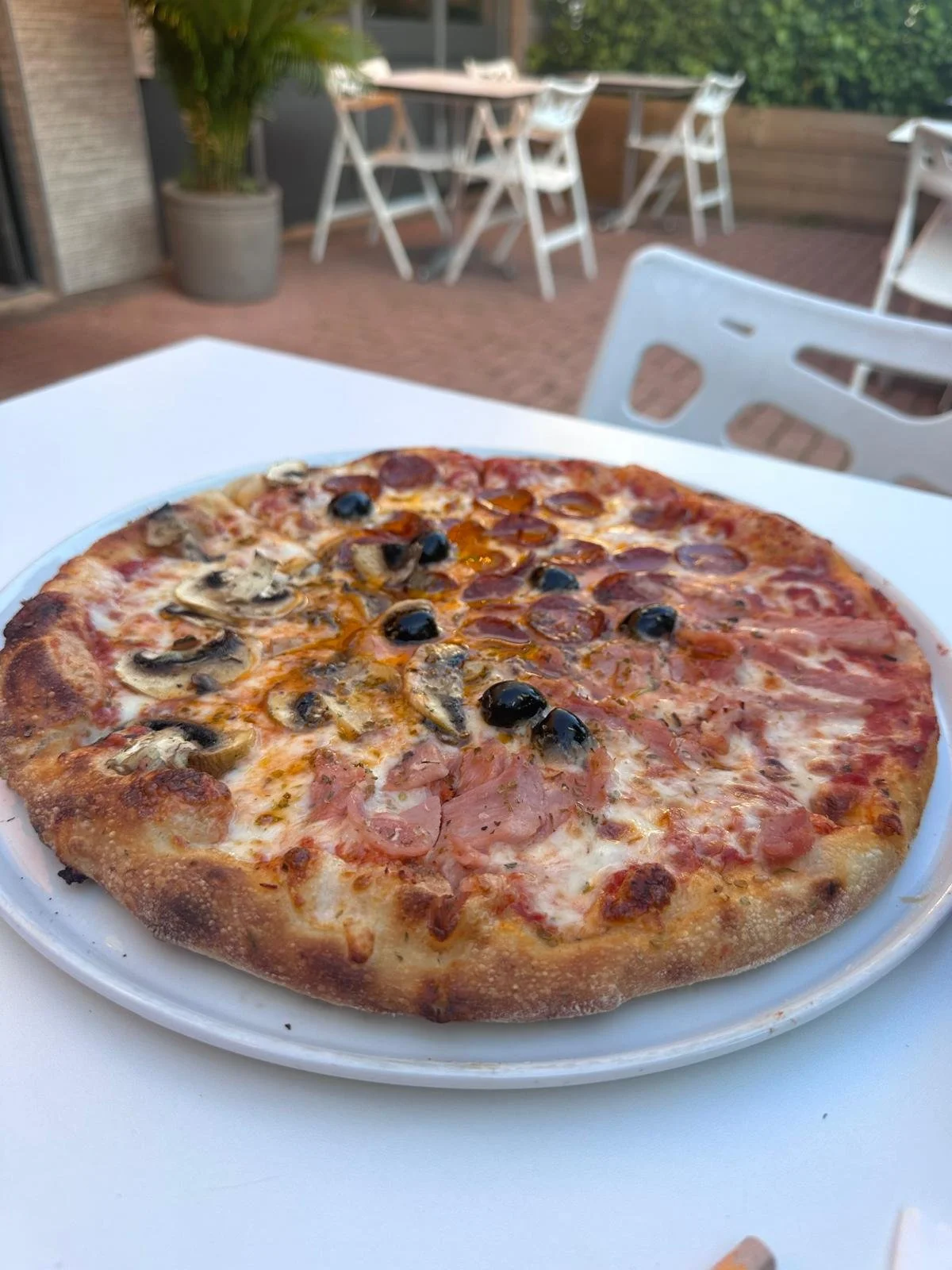 Close-up of a half mushroom, half pepperoni pizza on a white plate, outdoor patio with tables and chairs in the background.