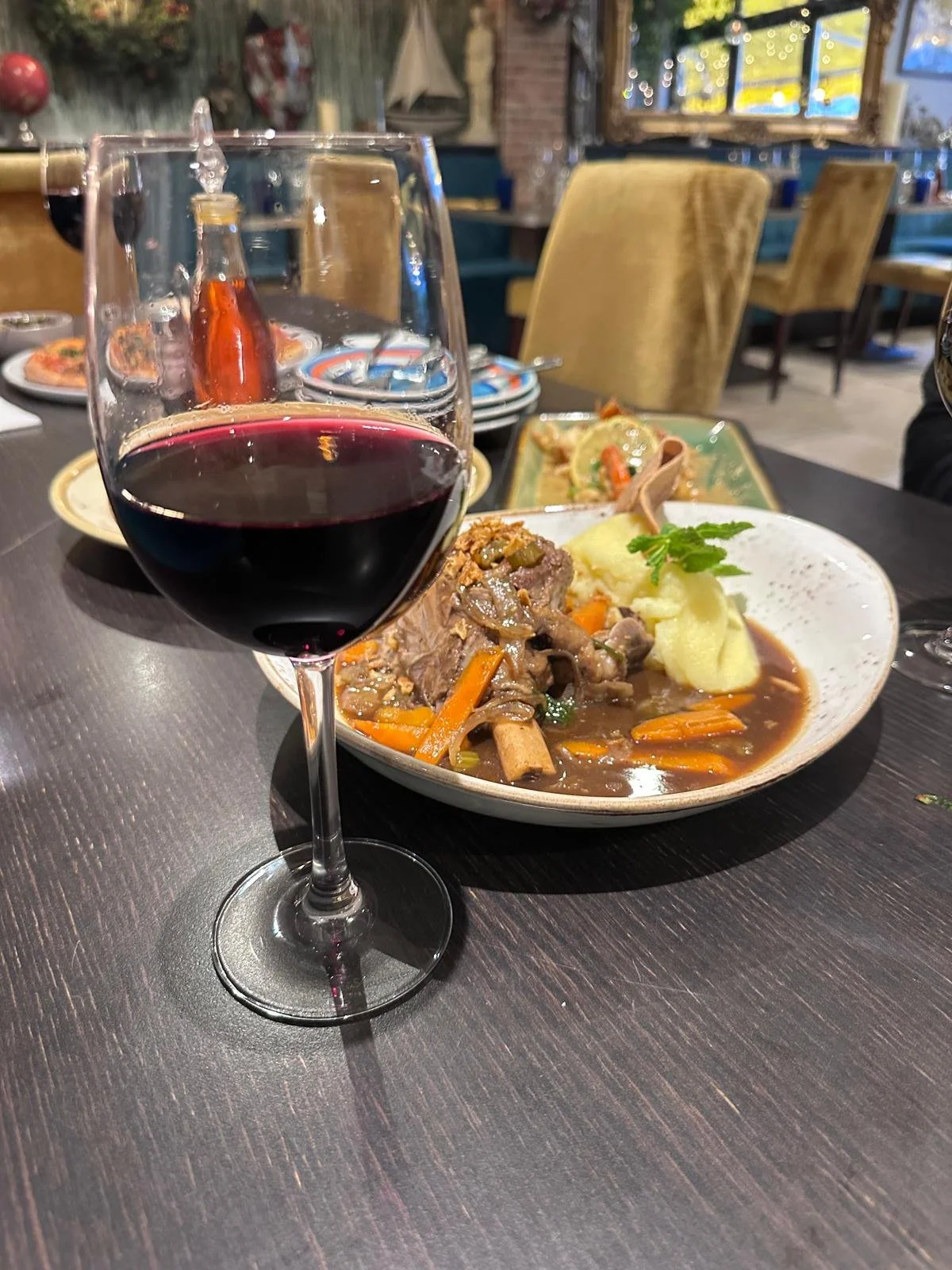 A glass of red wine and a plate of beef stew with mashed potatoes on a dark wooden dining table in a restaurant with yellow chairs and decorative lights.