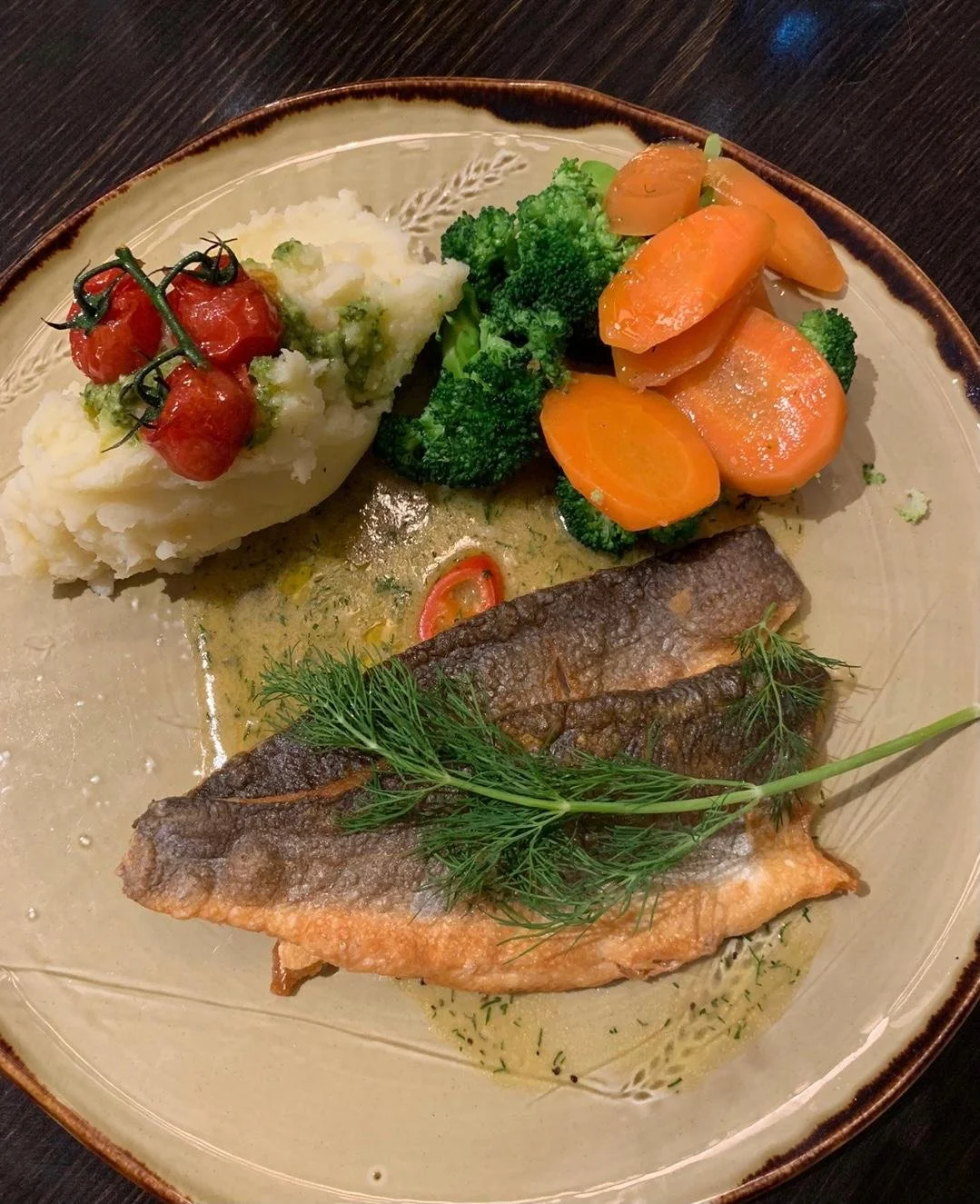Plate with mashed potatoes topped with cherry tomatoes, cooked broccoli, sliced carrots, and a piece of grilled fish garnished with dill.