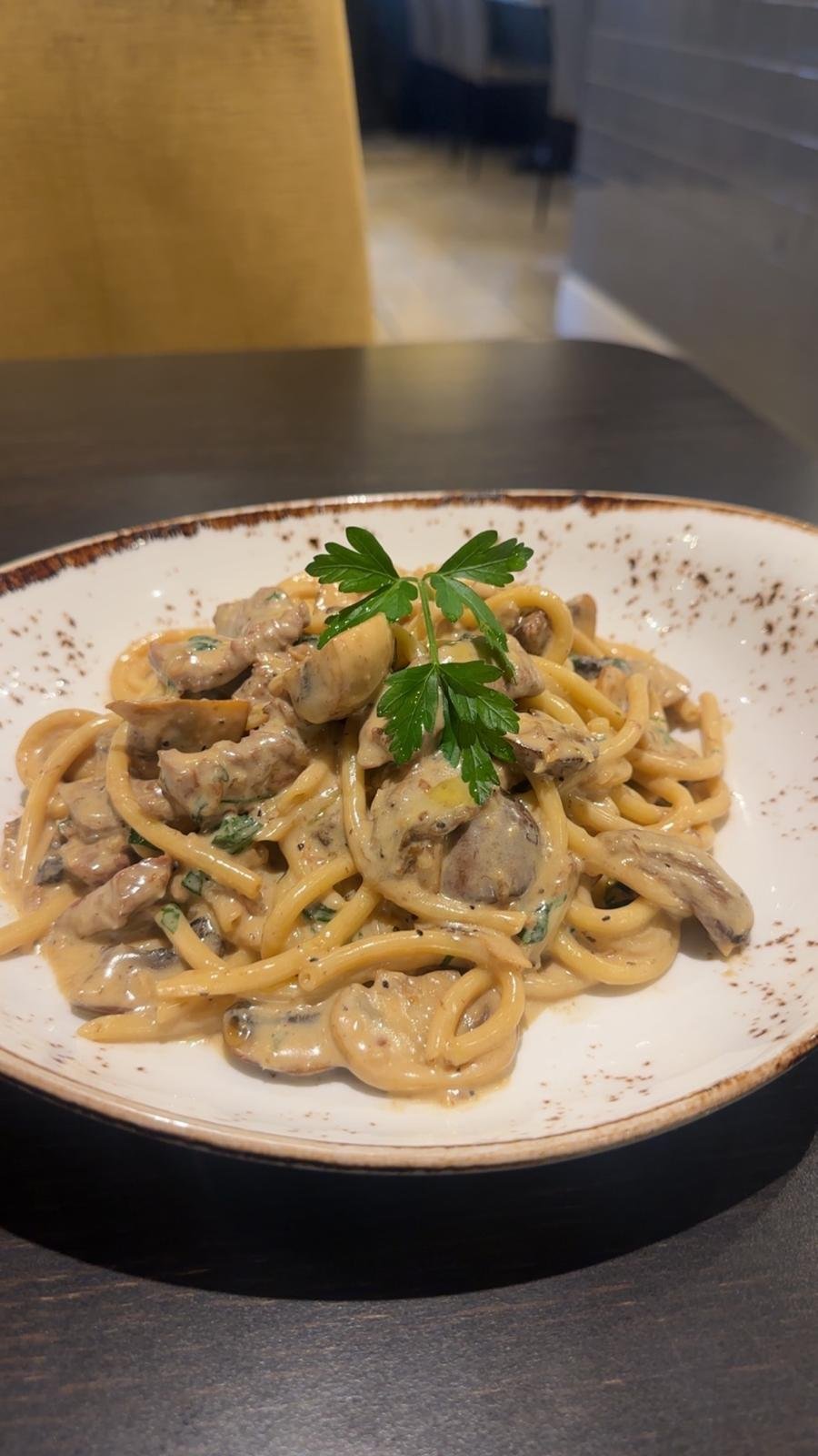 A bowl of cream pasta with mushrooms, garnished with parsley on top, on a dark table in a restaurant setting.