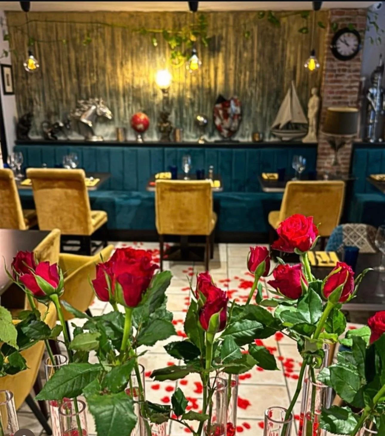 The image shows a decorated dining room with pink roses in glass vases in the foreground on a table. The room features yellow chairs around a dark dining table, and the background has a blue paneled wall with decorative items such as sculptures and a sailboat. There are hanging lights and a clock on the brick wall.
