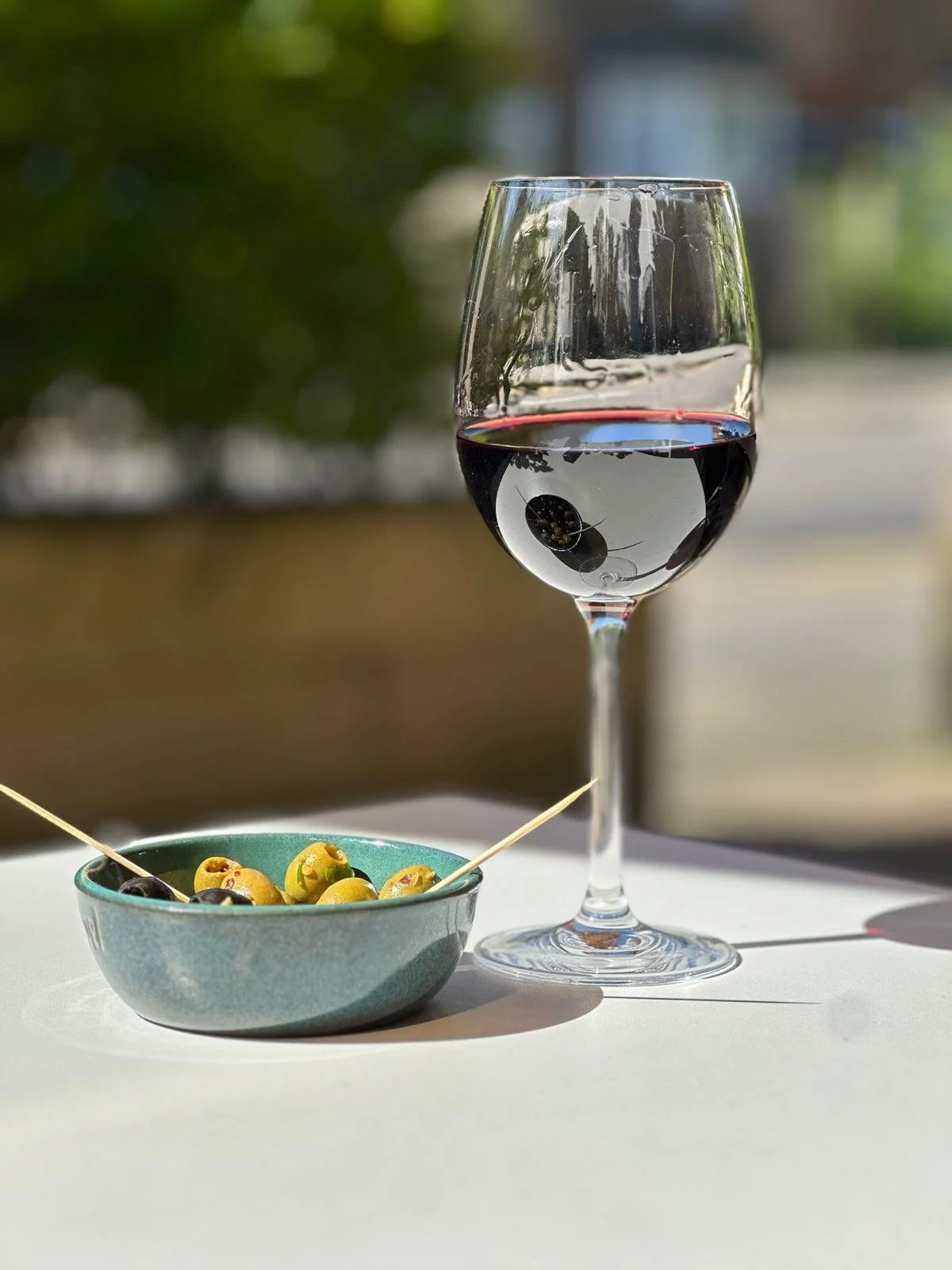 A glass of red wine and a small bowl of green olives on a white surface outdoors with a blurred background of trees and a building.