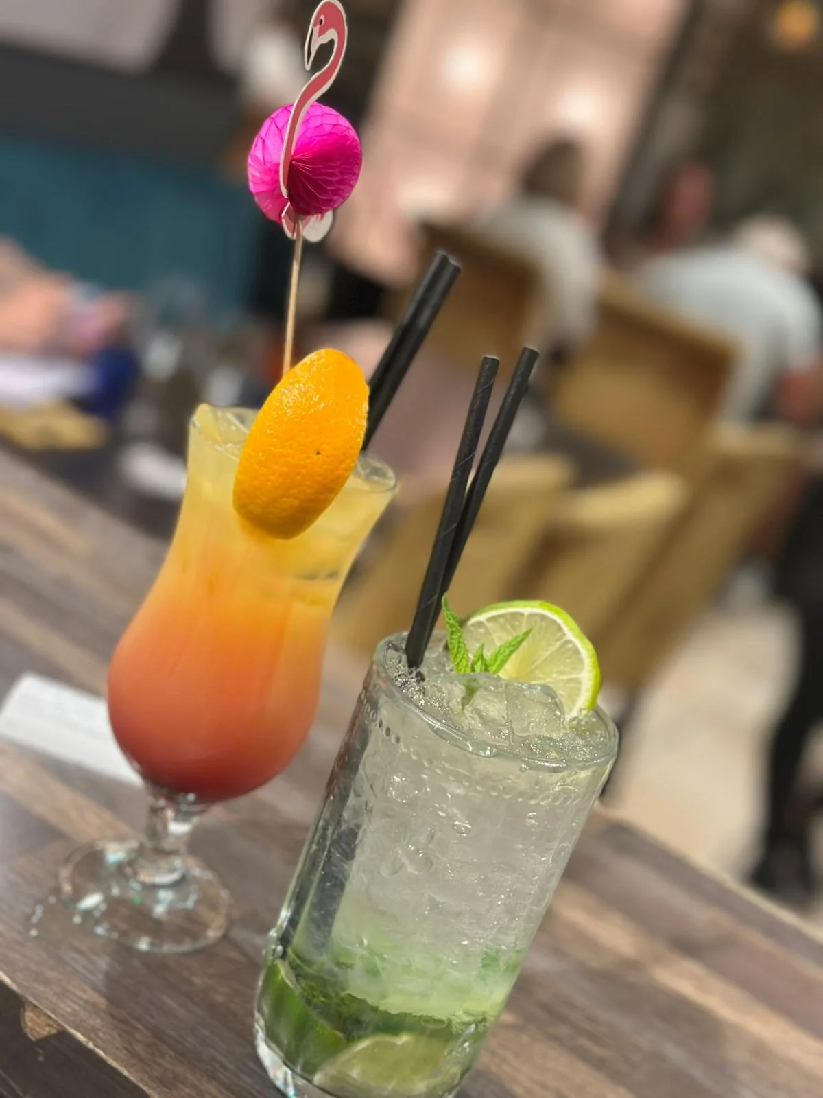Two colorful cocktails with garnishes on a wooden table, one with a pink paper umbrella and orange slice, the other with lime slices and mint leaves.