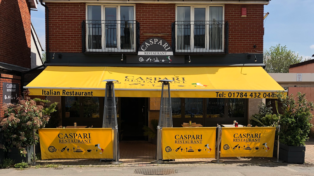 Front of an Italian restaurant called Caspari with a yellow awning and outdoor seating area; the restaurant's name and contact number are on the awning, and there are decorative signs and plants around the seating area.