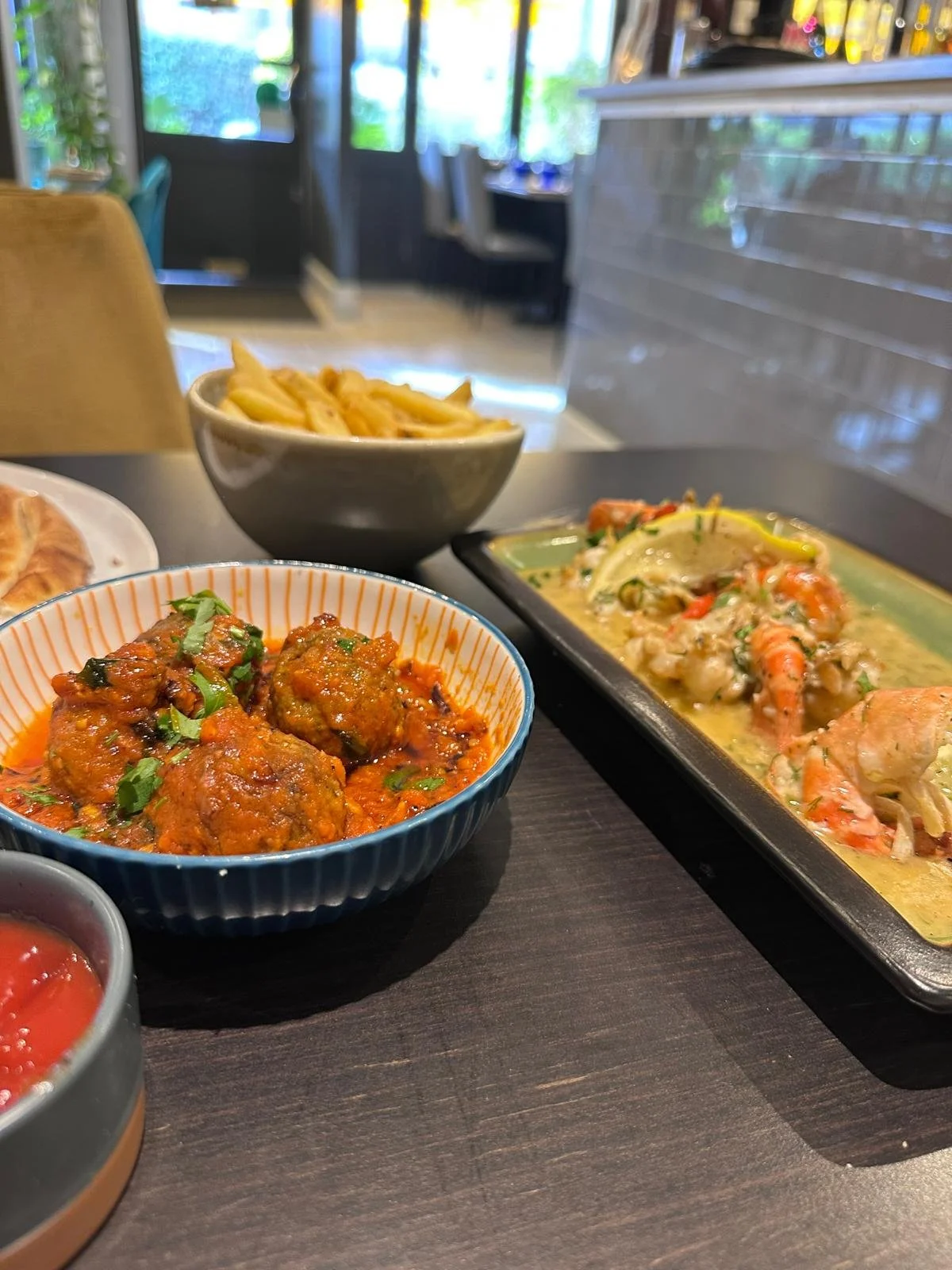 Assorted Indian dishes on a dark wooden table, including a bowl of meatballs in red sauce, lentil soup with vegetables, and a vegetable curry with shrimp, in a restaurant setting.
