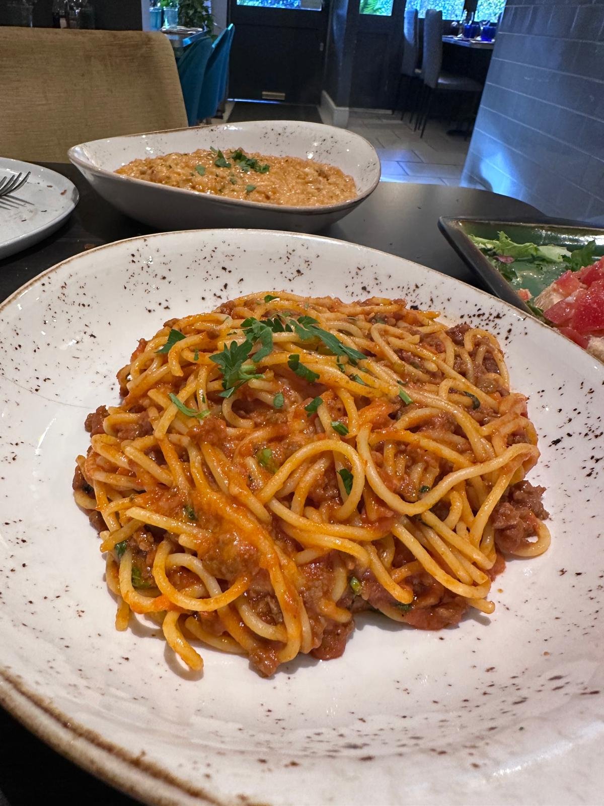 Close-up of a plate of spaghetti with meat sauce garnished with fresh herbs, on a white speckled ceramic plate. In the background, a dish of lasagna and a salad are visible on a dining table in a restaurant setting.